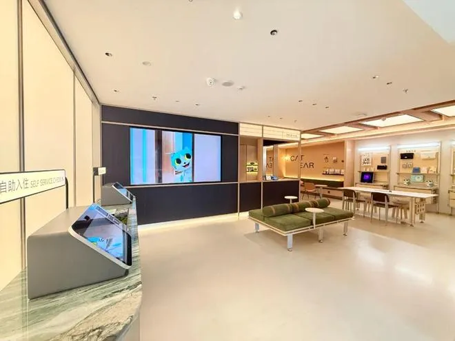 成都猫耳朵酒店大堂现场：左侧是自动办理入住的智能自助机（屏幕朝向住客），中部背景墙投影出 CAT EAR HOTEL 标识与青猫 IP 形象，右侧是品牌文创展示区与高脚桌椅